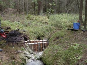 Vaihtoehtoinen teksti: Kuvassa on lähde ja lähteestä kaivettuun uomaan puutavarasta tehty patorakenne.