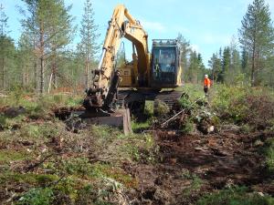 En gul grävmaskin lägger igen ett dike. En person står lite längre bort.