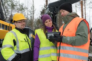 Metsäammattilaiset tarkastelevat tietoja tabletilta metsäkoneen vieressä metsässä.