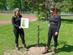 Två personer står bredvid en nyplanterad trädplanta. Den ena hålkler ett diplom.