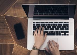 Händer på en bärbar dators tangentbord. En telfon ligger på bordet bredvid datorn.