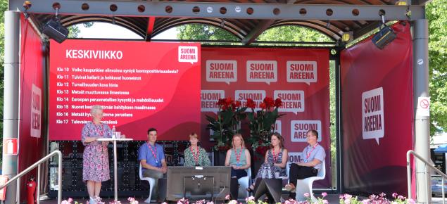 Juontaja ja paneelikeskustelijat SuomiAreenan lavalla.