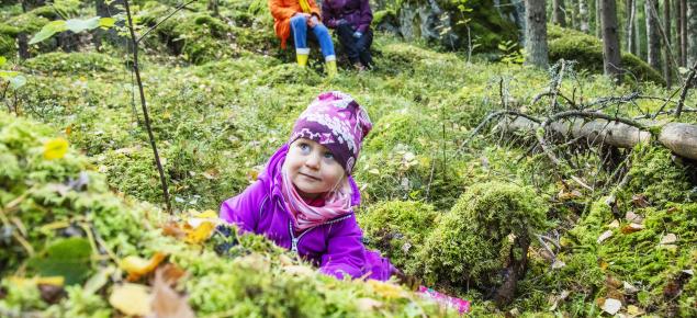 Ett litet barn sitter på marken i skogen och tittar uppåt. Bakom barnet sitter två vuxna på en sten i skogen.