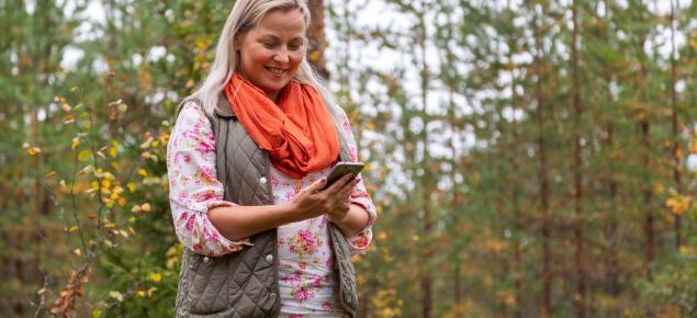 En person tittar på en mobiltelefon och ler medan hen går i skogen.