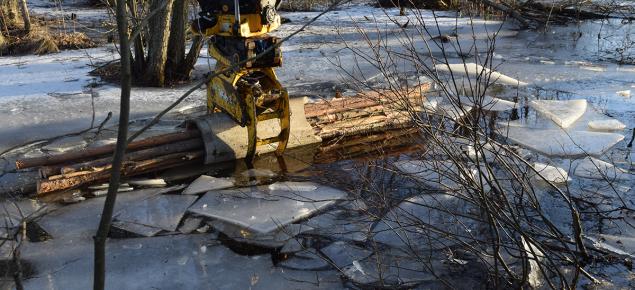 Kaivinkoneen koura laittaa puunippua veteen talvella. 