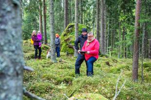 Människor i olika åldrar och en hund i skogen, två personer tittar på en surfplatta.