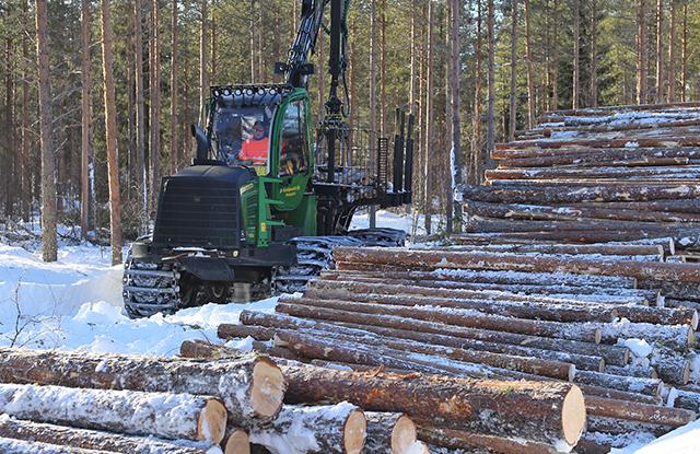 Vihreä metsäkone puupinon takana talvisessa maisemassa.