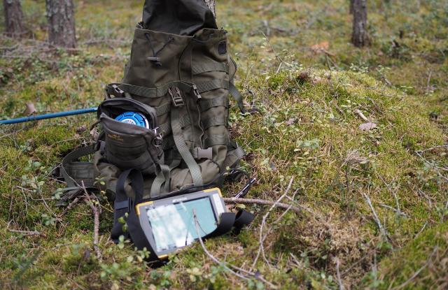 Reppu ja mittausvälineitä metsässä nojaamassa puuhun.